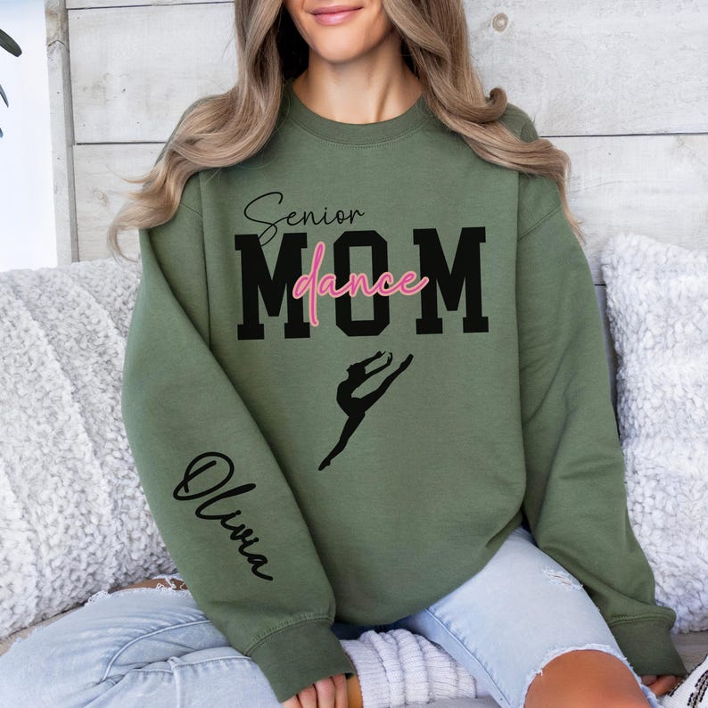 May include: A green sweatshirt with the text "Senior Dance Mom" in black and pink lettering. A silhouette of a dancer is below the text. The word "Olivia" is written in cursive on the left sleeve.