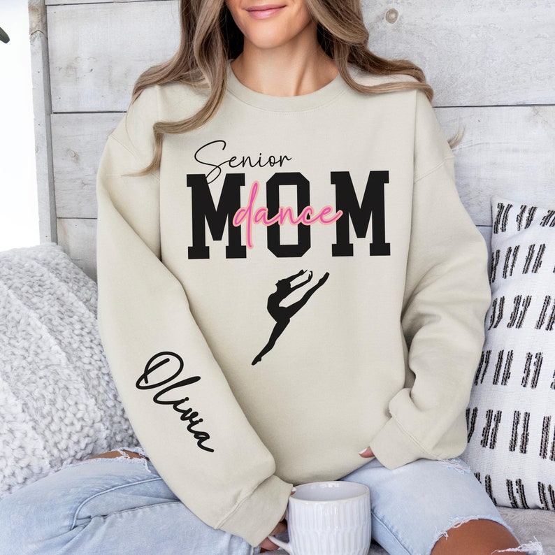 May include: A beige sweatshirt with the text "Senior Dance Mom" in black with a pink "dance" and a silhouette of a dancer. The left sleeve has the name "Olivia" written in black.