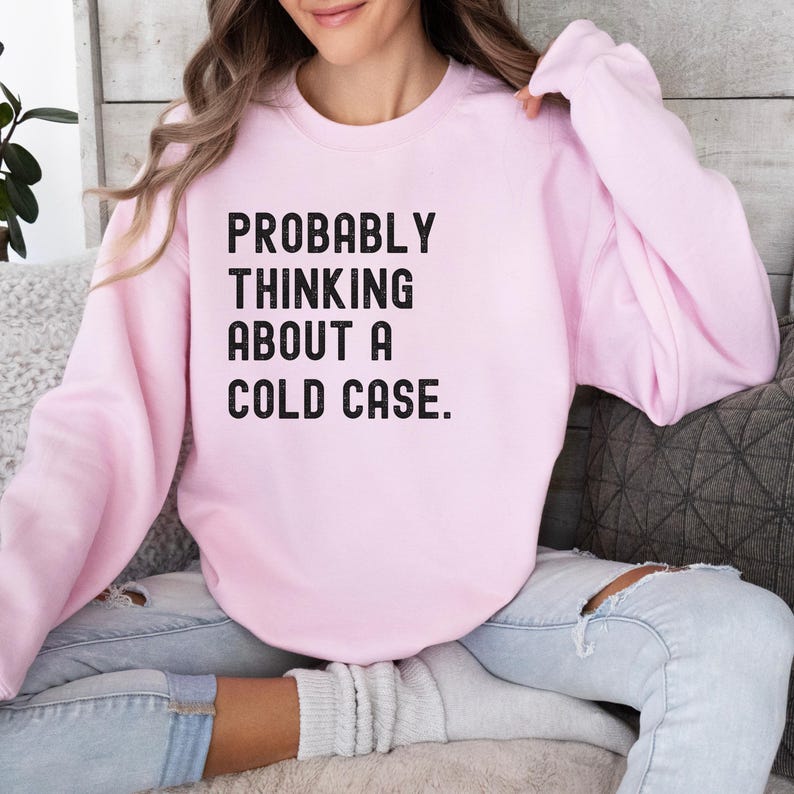 May include: A pink sweatshirt with the text "PROBABLY THINKING ABOUT A COLD CASE." printed in black.