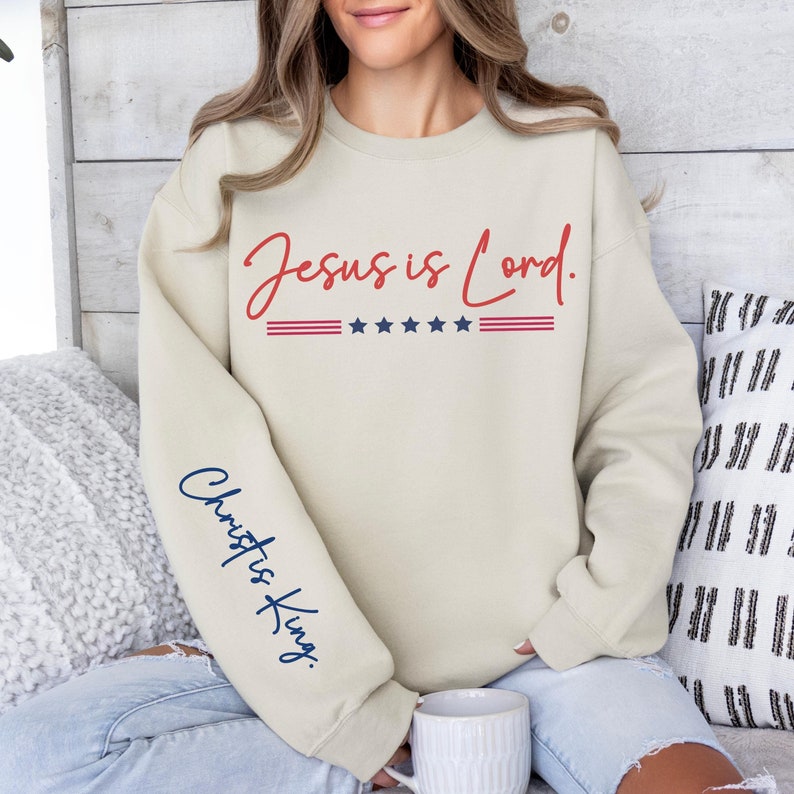 May include: A beige sweatshirt with the text "Jesus is Lord." in red script with three red stripes and five blue stars above the text. The left sleeve has the text "Christ is King." in blue script.