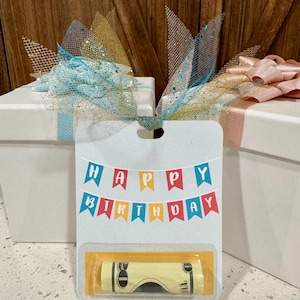 May include: A white gift box with a blue ribbon and a decorative bow. A gift tag with the words "HAPPY BIRTHDAY" in colorful banner letters is attached. A rolled-up bill is visible in a clear plastic sleeve.