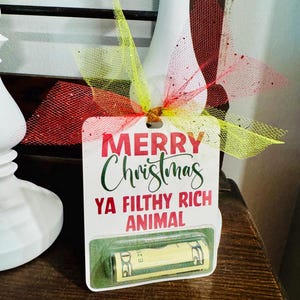 May include: A Christmas gift tag with the text "MERRY Christmas YA FILTHY RICH ANIMAL" in red and green. The tag is decorated with a red and yellow bow and contains a rolled-up twenty-dollar bill.