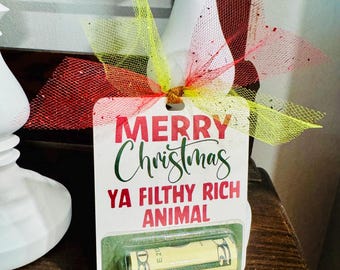 May include: A Christmas gift tag with the text "MERRY Christmas YA FILTHY RICH ANIMAL" in red and green. The tag is decorated with a red and yellow bow and contains a rolled-up twenty-dollar bill.
