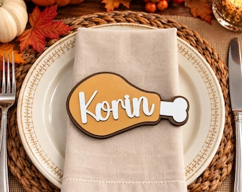 May include: A tan napkin ring shaped like a chicken leg with a dark brown outline and a white bone end. The name Korin is written in white cursive script across the center of the tan portion of the chicken leg shaped napkin ring.