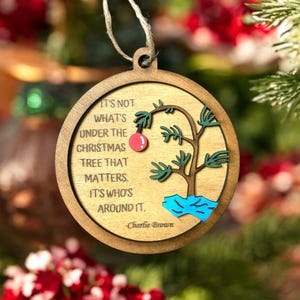 May include: A round wooden Christmas ornament with a quote from Charlie Brown: "It's not what's under the Christmas tree that matters. It's who's around it." The ornament features a cartoon depiction of a small, lopsided Christmas tree with a red ornament.