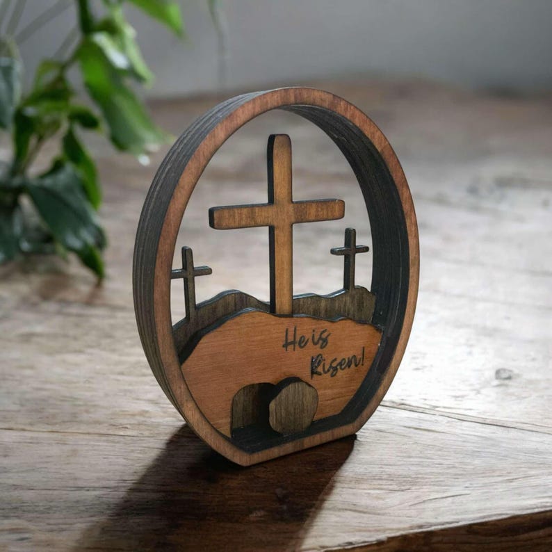 May include: Wooden Easter cross decoration with a brown finish. The cross is surrounded by a circle with a cutout of a hill and a tomb. The text "He is Risen!" is engraved on the base.