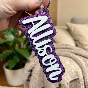 May include: A purple glitter keychain with the name "Allison" in white script. The keychain is attached to a silver metal loop. The background includes a green plant in a white pot and a beige blanket.