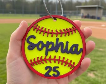 Peut inclure: Une étiquette ronde sur le thème du softball avec un centre jaune, une bordure rouge et des détails de couture rouges. Le nom "Sophia" est en écriture bleu foncé, et le numéro "25" est en dessous. Une boucle en fil d'argent est attachée en haut.