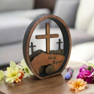 May include: Wooden Easter cross and tomb decoration with the text "He is Risen!"  The decoration is brown and features a cross with a tomb at the base.