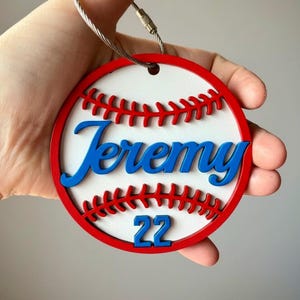 May include: A baseball-themed keychain with a red and white design. The keychain features the name "Jeremy" in blue script and the number "22" in blue. Red stitching detail and a red border complete the design.