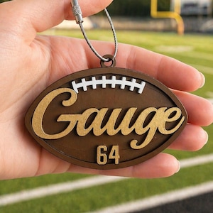 May include: An oval-shaped brown wooden ornament shaped like a football, featuring the gold script text Gauge, the number 64 in gold, and white stitched detailing. A silver metal wire loop is attached to the top for hanging purposes.