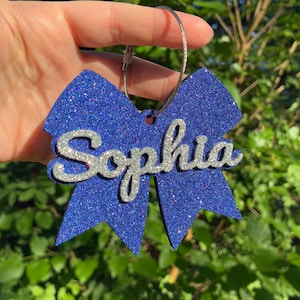 May include: A blue glitter bow-shaped keychain featuring the name Sophia in silver glitter script lettering. The ornament is attached to a thin, circular metal wire loop, creating a decorative accessory with a sparkling finish.