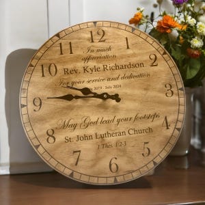 Puede incluir: Un reloj de madera con números romanos y una aguja negra. La esfera del reloj tiene una inscripción que dice: "In much appreciation Rev. Kyle Richardson For your service and dedication July 2019 - July 2024 May God lead your footsteps St. John Lutheran Church 1 Thes. 1:2-3"