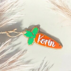 Peut inclure: Une d&eacute;coration en bois en forme de carotte avec un dessus vert feuillu et le nom "Korin" &eacute;crit en blanc sur la carotte orange.