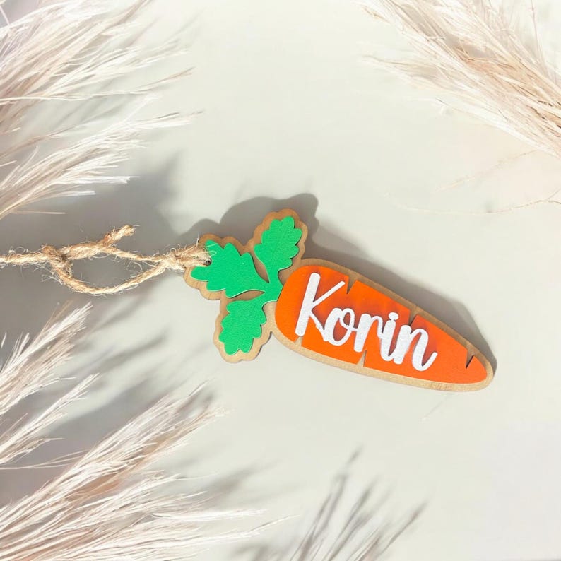 Peut inclure: Une d&eacute;coration en bois en forme de carotte avec un dessus vert feuillu et le nom "Korin" &eacute;crit en blanc sur la carotte orange.