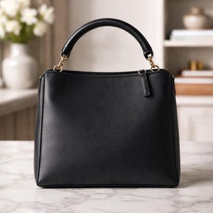 May include: A black handbag with a rounded handle and gold-colored hardware. The structured bag has a rectangular shape with a top zipper closure and a small, attached leather tab. The bag is sitting on a marble surface.