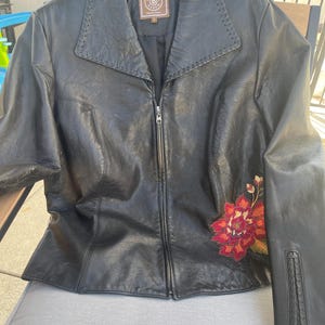 May include: Black leather jacket with a floral embroidery on the right side. The jacket has a zipper closure and a collar. The label on the jacket reads "Douglas Ranch" with a "D" logo.