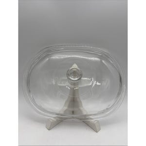 May include: A clear, oval-shaped glass lid featuring a smooth, transparent surface and a centered, circular knob handle. The lid has a raised outer rim designed to fit securely onto a corresponding container, showcasing a simple, functional design.