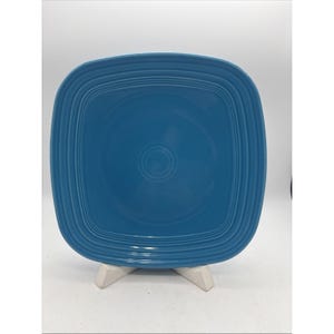 May include: A square ceramic plate in a solid medium blue color. The plate features a series of concentric embossed rings around the perimeter and a single circular ring embossed in the center of the flat surface.