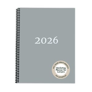 Pode incluir: Um planejador cinza de 2026 com encadernação em espiral preta. A capa apresenta o ano em texto branco e um adesivo circular que diz "Marketing Planner for Lawyers" e "Put Your Firm's Growth on Autopilot."