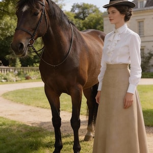 May include: A woman in a white blouse with ruffles and a long tan skirt stands next to a brown horse. She wears a black hat. The horse has a bridle and is standing on a grassy area near a path.