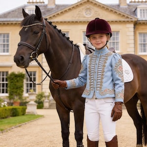 May include: A young equestrian stands next to a dark brown horse. The equestrian wears a light blue jacket with gold trim, white riding pants, a burgundy helmet, and brown gloves and boots. The horse has a bridle and is standing on a paved path.
