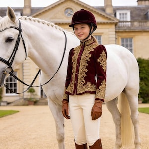 May include: A young person in equestrian attire stands next to a white horse. The person wears a burgundy jacket with gold embroidery, a matching helmet, and white pants. The horse has a black bridle. The background features a large building.