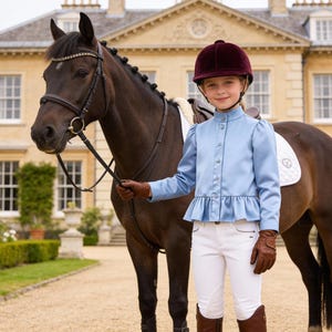 May include: A young equestrian stands beside a dark brown horse. The rider wears a light blue riding jacket, white breeches, brown boots, gloves, and a burgundy helmet. The horse has a bridle and saddle. The background features a large building and a gravel driveway.