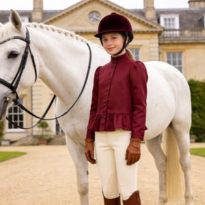 May include: A young person in equestrian attire stands next to a white horse. They wear a burgundy riding jacket, helmet, tan gloves, and white breeches. The horse has a black bridle. The background features a large building with many windows.