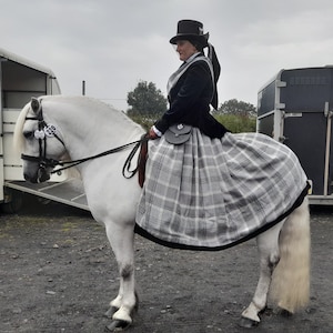 May include: A woman in a black top hat and a plaid skirt rides a white horse. The horse is wearing a bridle and a saddle. The woman is holding the reins in her hands.