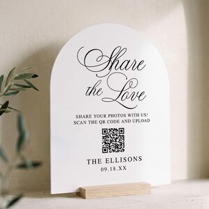 May include: An arched white sign with black text reading Share the Love, SHARE YOUR PHOTOS WITH US! SCAN THE QR CODE AND UPLOAD, THE ELLISONS, and 09.18.XX. The sign features a central QR code and is supported by a rectangular light wood base.
