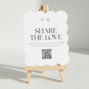 May include: A white sign with a scalloped edge, displayed on a small wooden easel. The sign reads "SHARE THE LOVE" in a bold font, with a QR code below. The initials "E | M" are at the top.