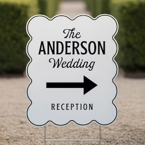 May include: A white wedding reception sign with a scalloped edge. The sign reads "The Anderson Wedding" with an arrow pointing right to "Reception" in black text. The sign is mounted on a metal stake.