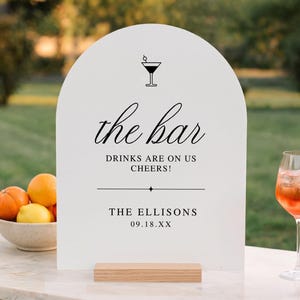 May include: An arched white sign with black text reading the bar, DRINKS ARE ON US CHEERS!, THE ELLISONS, and 09.18.XX. The sign features a martini glass icon and a horizontal line, and it is supported by a rectangular light wood base.