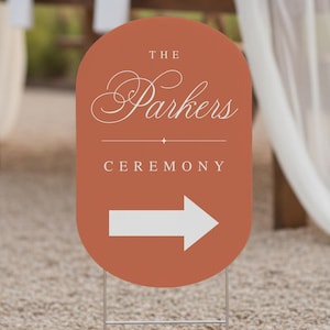 May include: An oval-shaped sign in a terracotta color, with the words "THE Parkers CEREMONY" in white script and a white arrow pointing right. The sign is mounted on a metal stake, suitable for outdoor use at a wedding or event.