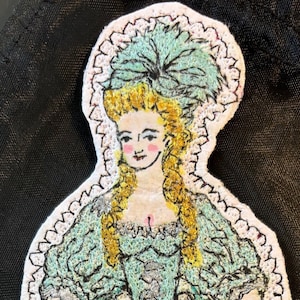 Puede incluir: Parche bordado de una mujer con un vestido turquesa con ribete de encaje blanco. Tiene el pelo rubio y lleva un sombrero turquesa con una pluma. El vestido está decorado con flores rosas.