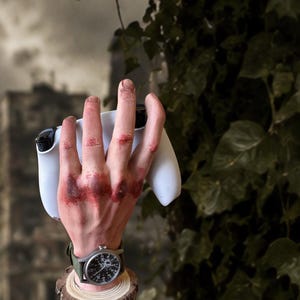 Puede incluir: Una escultura realista de una mano humana con nudillos magullados de tono rojizo que sostiene un mando de videojuego blanco. La mano lleva un reloj de pulsera con correa verde oscuro y esfera agrietada, montado sobre una base de madera.