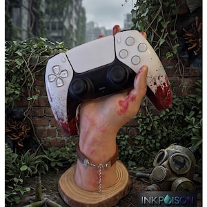 Peut inclure: Une manette de jeu blanche avec des éclaboussures rouges semblables à du sang, tenue par une sculpture de main réaliste. La main porte un bracelet en cuir marron avec une chaîne argentée. La base est une tranche de bois, logo INKPOISON.