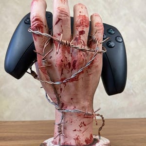 May include: A realistic sculpture of a pale, blood-stained hand standing upright on a circular wooden base. The hand is wrapped in metallic barbed wire and is positioned to hold a black video game controller behind its fingers.