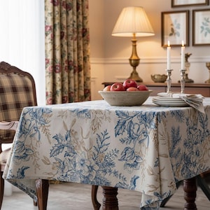 May include: A cream-colored tablecloth with a blue floral pattern covers a wooden table. The table is set with a bowl of red apples, plates, and candles. A plaid chair and floral curtains are in the background.