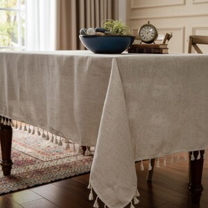 Op de afbeelding: Een beige rechthoekig tafelkleed met een franje van kleine kwastjes langs de randen. Het tafelkleed is gedrapeerd over een donkere houten tafel. Een blauwe kom met stenen en groen staat erop, samen met een kleine klok en boeken.