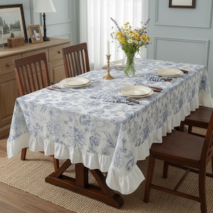 May include: A rectangular white tablecloth with a blue floral pattern and a ruffled edge. The table is set with plates, napkins, and silverware. A vase of flowers and a candle holder are on the table.