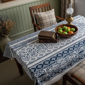 Puede incluir: Un mantel blanco con un estampado floral y geométrico azul cubre una mesa de madera. Un cuenco de madera lleno de manzanas verdes y naranjas está sobre la mesa, junto a una pila de libros. También se ven una silla de madera y una lámpara.