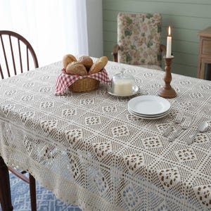 May include: A dining table set with a cream-coloured, openwork lace tablecloth. A basket of bread, butter dish, plates, silverware, and a lit candle in a wooden holder are arranged. A wooden chair is visible in the background.