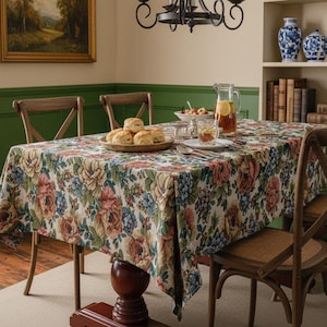 May include: A floral tapestry tablecloth with a cream background and a pattern of pink, blue, and yellow flowers. The table is set with a wooden plate of biscuits, glassware, and a pitcher of iced tea. Wooden chairs surround the table.