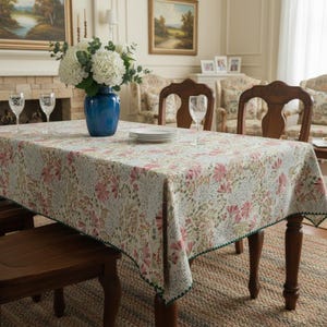 Puede incluir: Un mantel rectangular con un estampado floral en rosa, verde y beige cubre una mesa de madera. Un jarrón azul con flores blancas está en el centro. Copas de vino y platos están sobre la mesa.