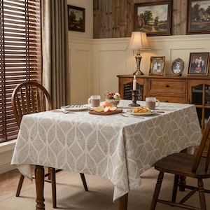 Puede incluir: Una mesa de comedor preparada para una comida. La mesa está cubierta con un mantel crema con un patrón geométrico repetido. Dos sillas de madera están colocadas a ambos lados de la mesa. La habitación tiene un ambiente cálido con muebles y decoración de madera.