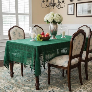May include: A dining room scene featuring a dark green lace tablecloth on a wooden table. The table is set with a silver vase of white flowers, a bowl of fruit, and candles. Four chairs with floral patterned upholstery surround the table.