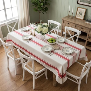 Puede incluir: Una mesa de comedor con un mantel blanco con rayas rojas. La mesa está puesta con platos, cubiertos y un centro de mesa floral con manzanas verdes. Sillas blancas con asientos de mimbre rodean la mesa, creando un ambiente acogedor.