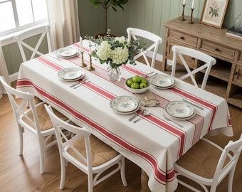 May include: A dining table set for a meal, featuring a white tablecloth with red stripes. The table is set with plates, silverware, and a centerpiece of flowers and green apples. White chairs with woven seats surround the table, creating a cozy dining atmosphere.